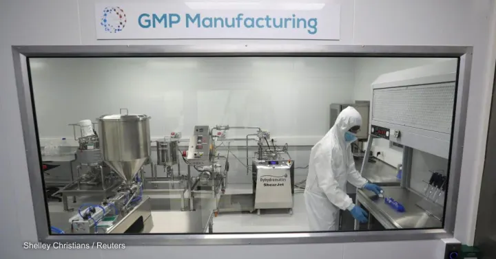 A technician in a white full-body suit works in a cleanroom labeled "GMP Manufacturing." The room, dedicated to local manufacturing in Africa, contains various stainless steel equipment and machinery.