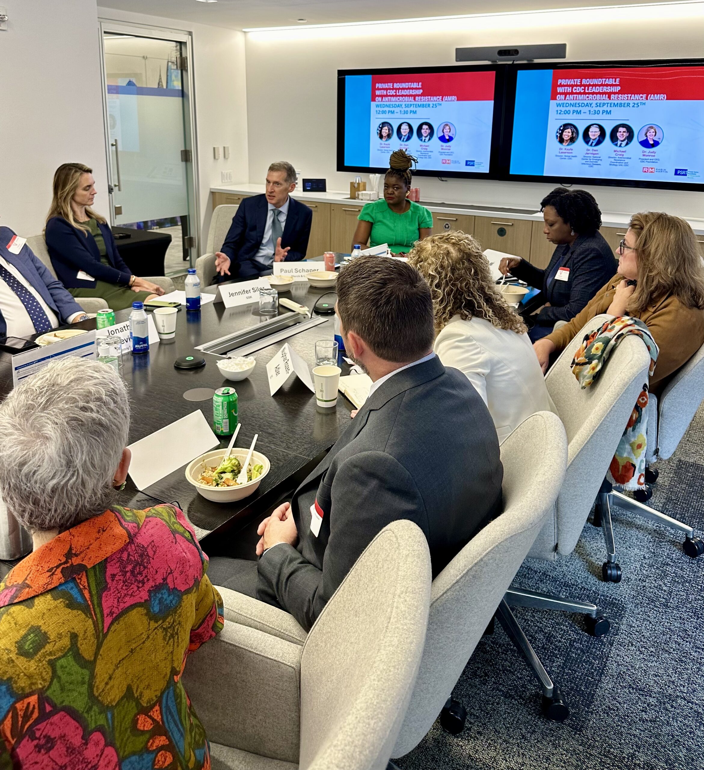 People sitting around a conference table during a meeting, discussing strategies to combat global antimicrobial resistance, with presentations displayed on screens in the background.