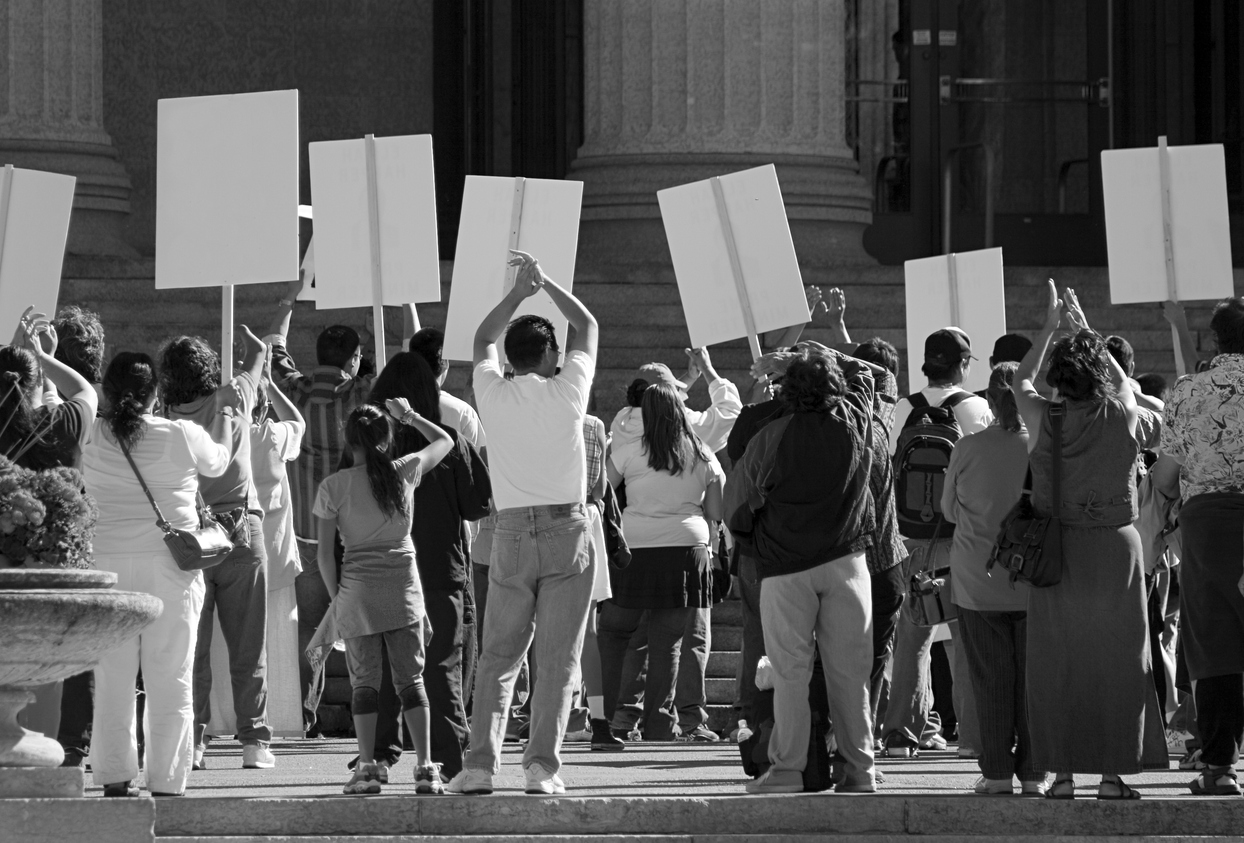 A crowd of people holding blank protest signs gather in front of a large building with columns, echoing the spirit of the Denver Principles as they advocate for health and empowerment.