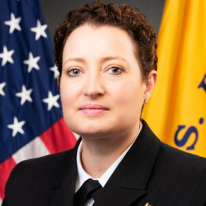 A woman in a military uniform standing in front of an American flag and a yellow flag.
