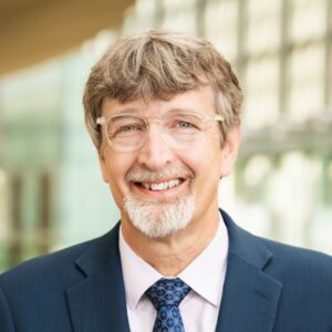 Headshot of a smiling man with a goatee and glasses, wearing a blue suit, light shirt, and patterned tie.