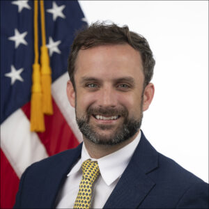 A man in a suit and yellow patterned tie smiles in front of an American flag.